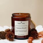 Candle jar labeled 'Autumn Woods' by Luca + Grey on a white surface with pinecones and nuts.