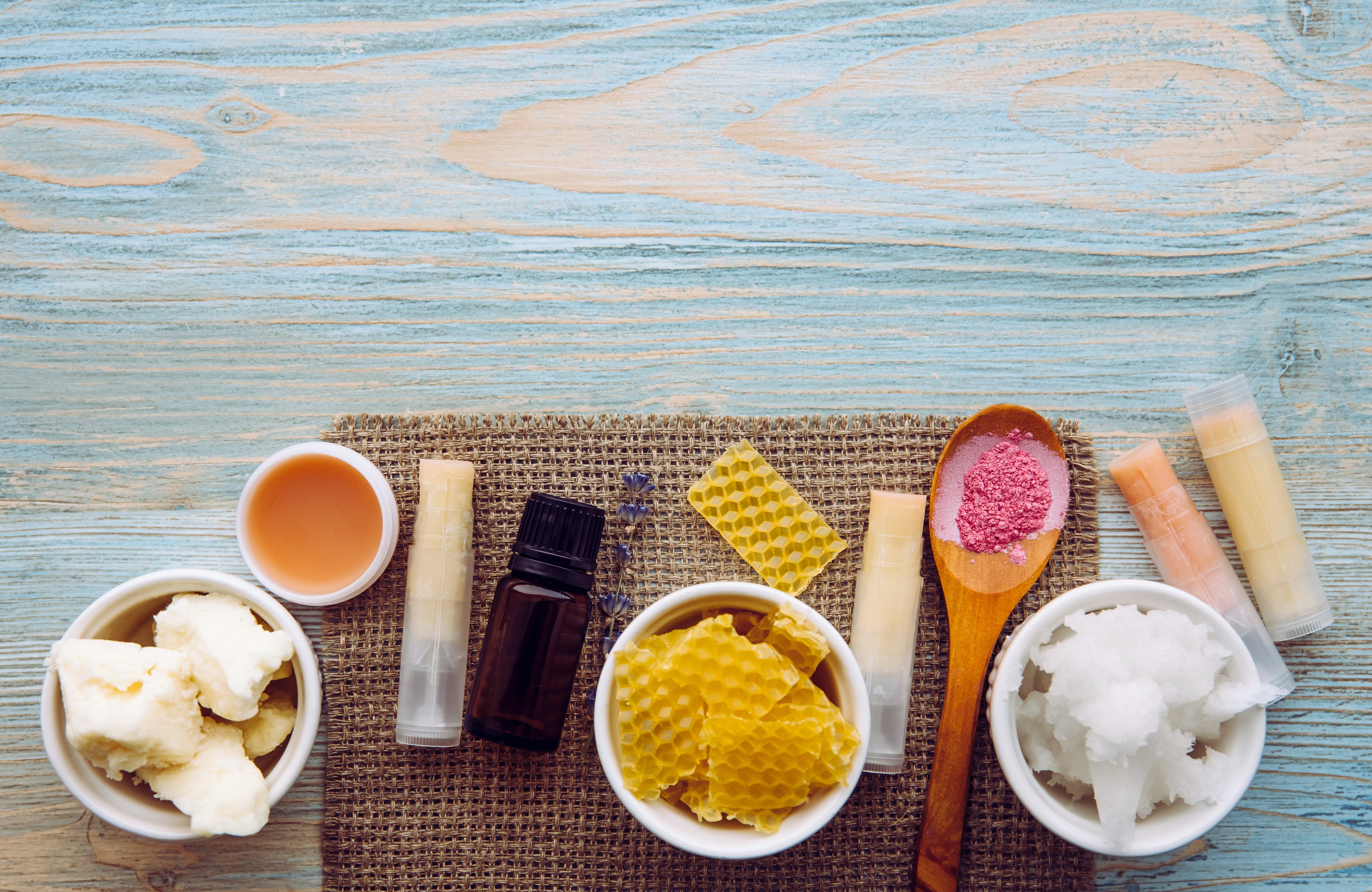 The image showcases natural beauty products on a blue wooden surface with a burlap cloth. Items include beeswax, honeycomb, essential oils, lip balms, shea butter, coconut oil, and pink salt, highlighting organic skincare ingredients.