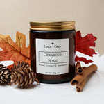 Candle labeled 'Cinnamon Spice' by Luca & Grey with autumn leaves and pinecones on a white surface.